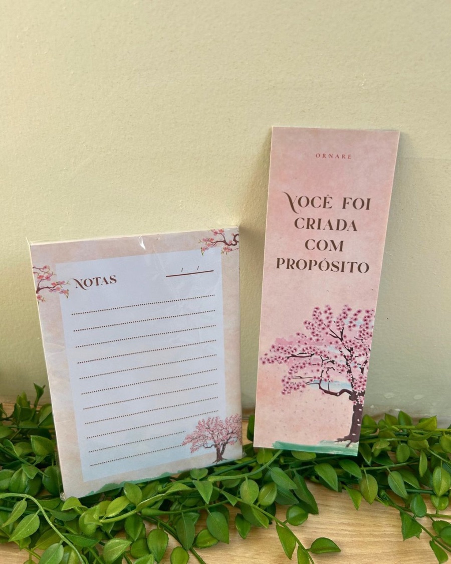 Bloco de notas e marca-página combinando, em tons de rosa, apoiados sobre folhagem.