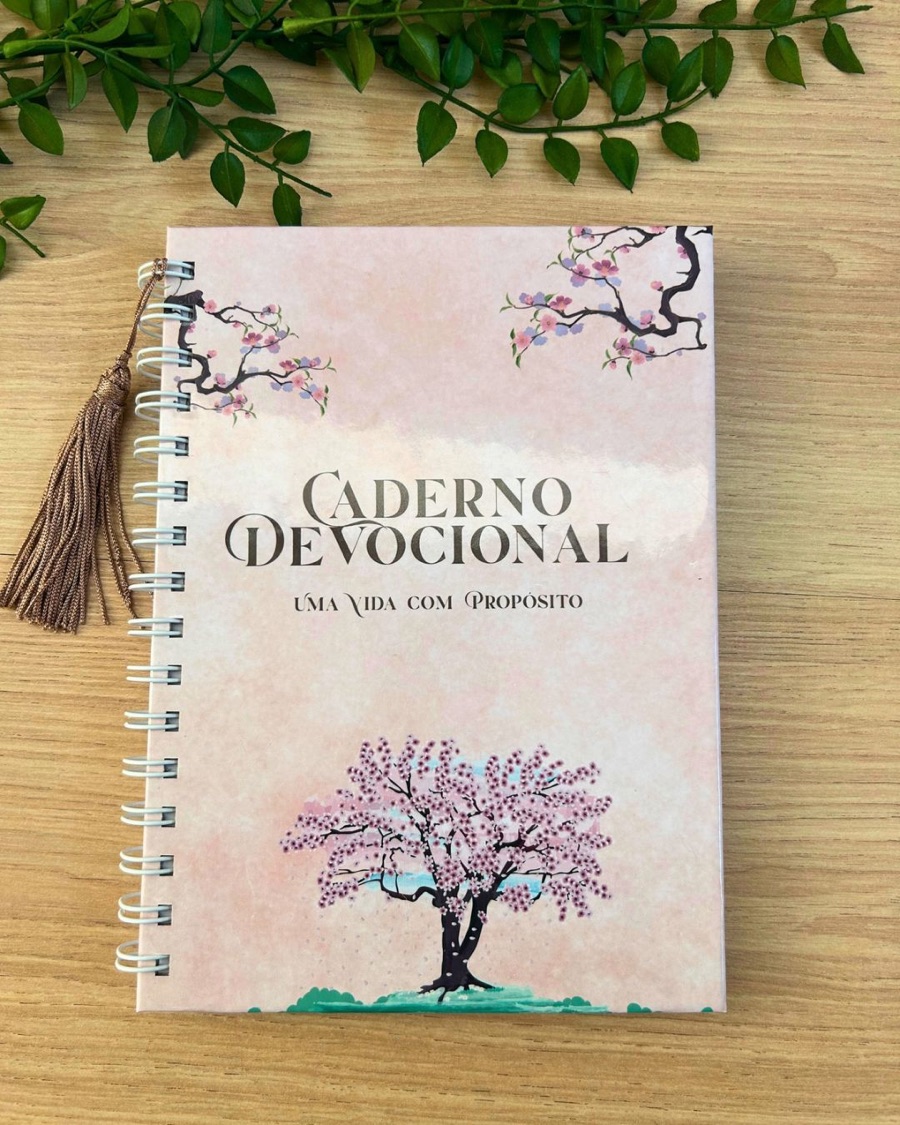 Caderno devocional com capa rosada, ilustração de cerejeira e pingente dourado, com espiral branca.