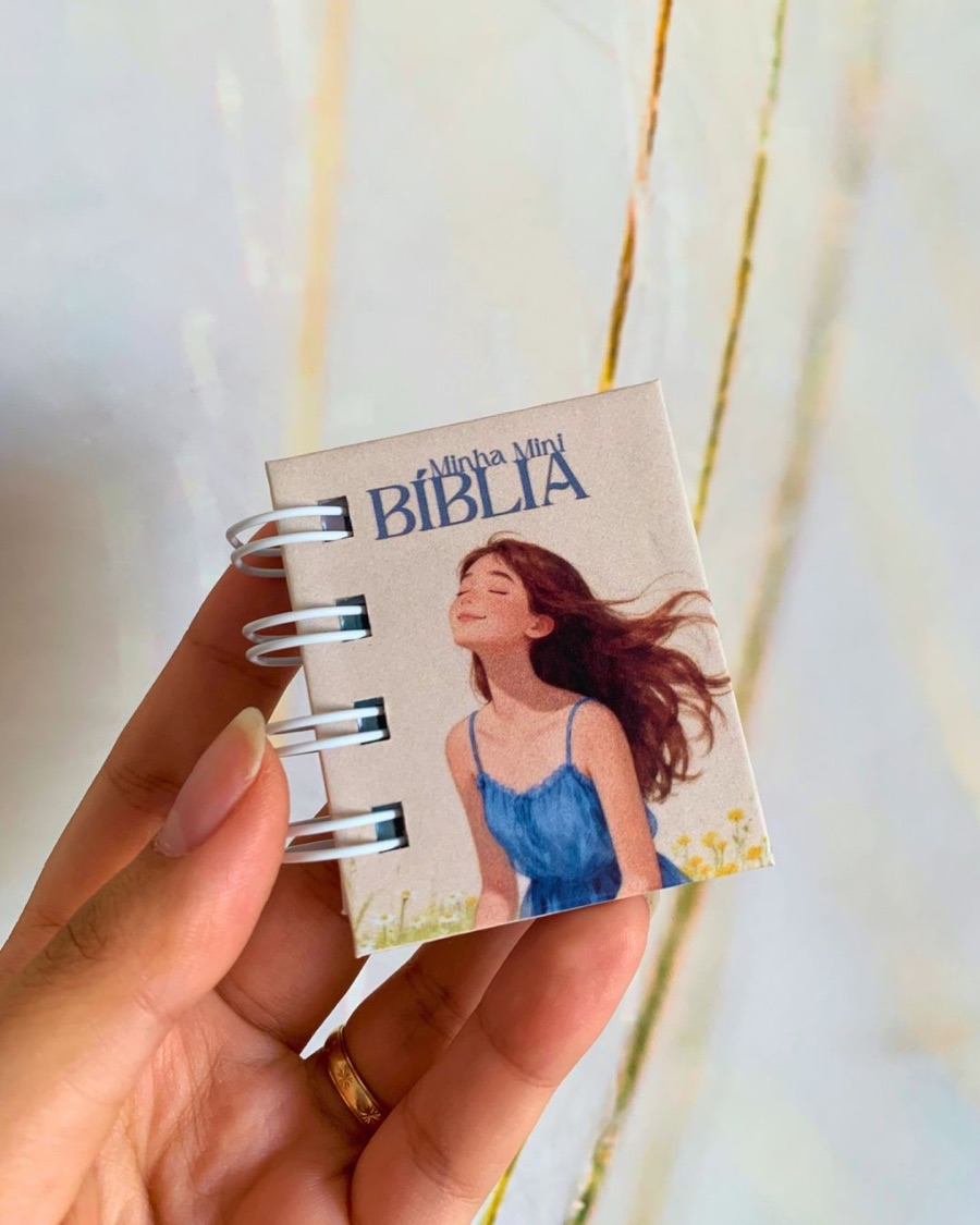 Mini Bíblia de bolso com capa ilustrada, presa por espiral, segurada na mão.